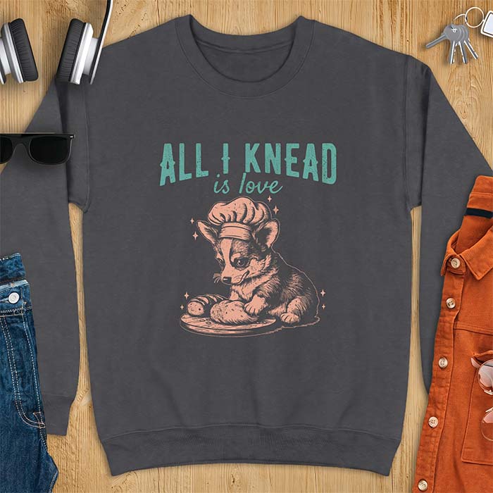 Dark Heather sweatshirt with a graphic of a corgi puppy wearing a chef's hat kneading a loaf of bread at a cutting board with sparkles around it, featuring the text All I Knead Is Love.