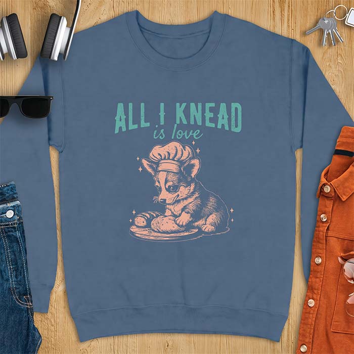 Indigo Blue sweatshirt with a graphic of a corgi puppy wearing a chef's hat kneading a loaf of bread at a cutting board with sparkles around it, featuring the text All I Knead Is Love.