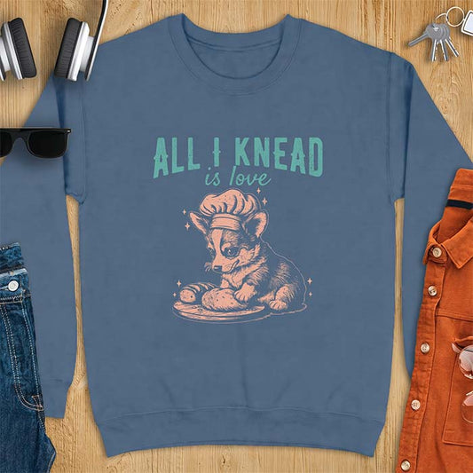 Indigo Blue sweatshirt with a graphic of a corgi puppy wearing a chef's hat kneading a loaf of bread at a cutting board with sparkles around it, featuring the text All I Knead Is Love.