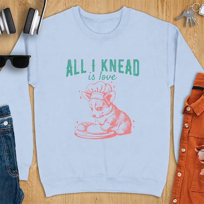 Light Blue sweatshirt with a graphic of a corgi puppy wearing a chef's hat kneading a loaf of bread at a cutting board with sparkles around it, featuring the text All I Knead Is Love.