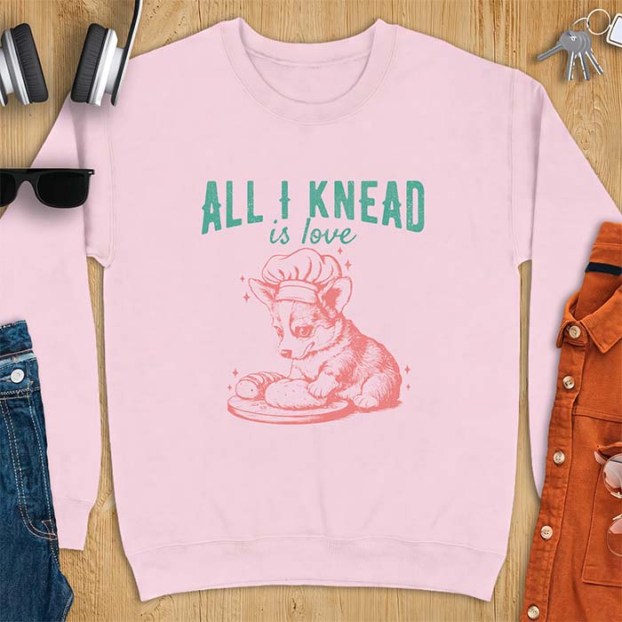 Light Pink sweatshirt with a graphic of a corgi puppy wearing a chef's hat kneading a loaf of bread at a cutting board with sparkles around it, featuring the text All I Knead Is Love.
