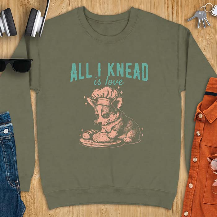 Military Green sweatshirt with a graphic of a corgi puppy wearing a chef's hat kneading a loaf of bread at a cutting board with sparkles around it, featuring the text All I Knead Is Love.