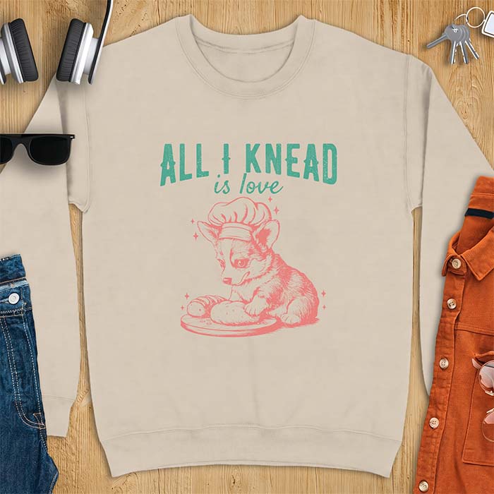 Sand sweatshirt with a graphic of a corgi puppy wearing a chef's hat kneading a loaf of bread at a cutting board with sparkles around it, featuring the text All I Knead Is Love.