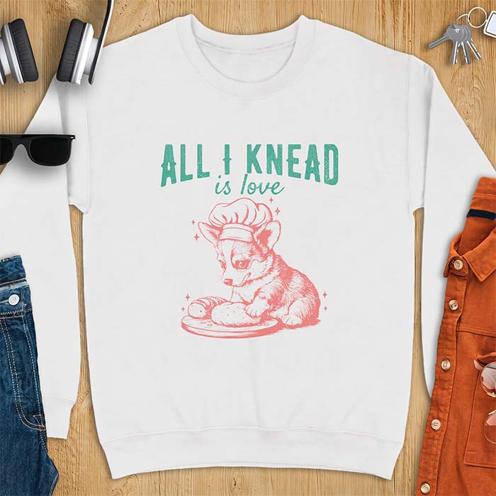 White sweatshirt with a graphic of a corgi puppy wearing a chef's hat kneading a loaf of bread at a cutting board with sparkles around it, featuring the text All I Knead Is Love.