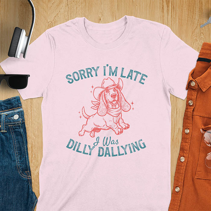 Light pink t-shirt featuring a galloping basset hound wearing a cowboy hat with long floppy ears flowing, text reads Sorry I'm Late I Was Dilly Dallying