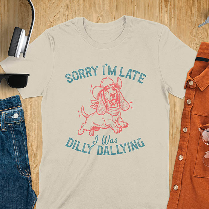 Sand color  t-shirt featuring a galloping basset hound wearing a cowboy hat with long floppy ears flowing, text reads Sorry I'm Late I Was Dilly Dallying