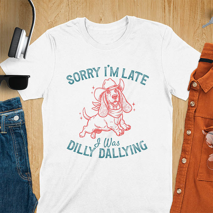 White t-shirt featuring a galloping basset hound wearing a cowboy hat with long floppy ears flowing, text reads Sorry I'm Late I Was Dilly Dallying