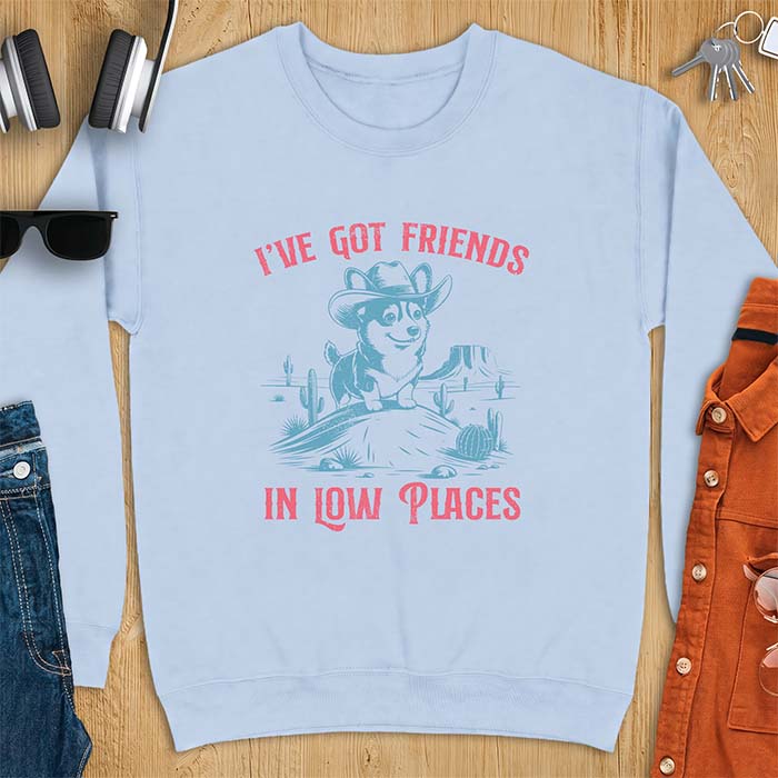 Light Blue sweatshirt with a western graphic of a corgi puppy in a cowboy hat standing on a desert rock surrounded by cacti and a mesa, with the text I've Got Friends In Low Places printed above and below it.