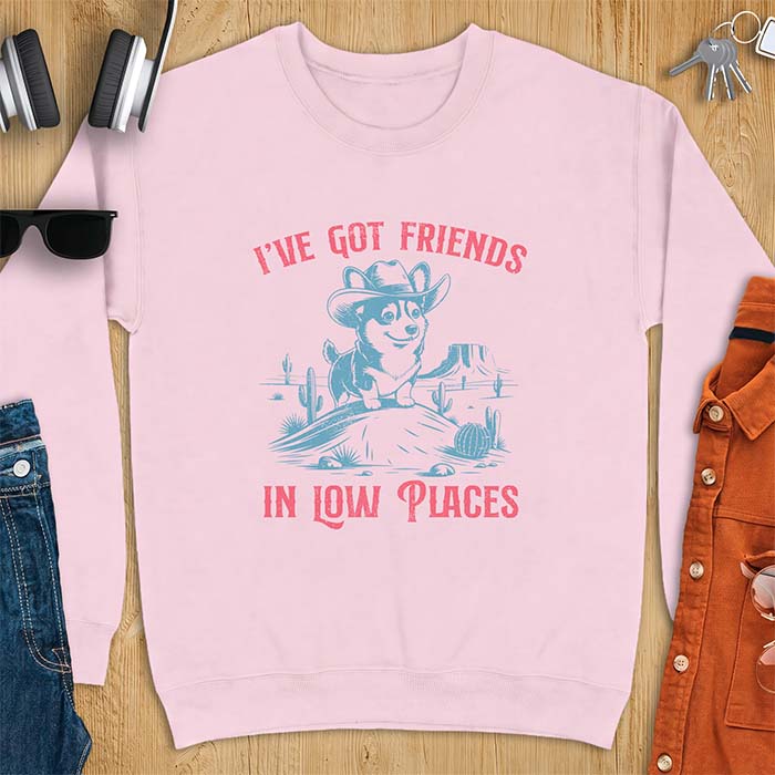 Light Pink sweatshirt with a western graphic of a corgi puppy in a cowboy hat standing on a desert rock surrounded by cacti and a mesa, with the text I've Got Friends In Low Places printed above and below it.