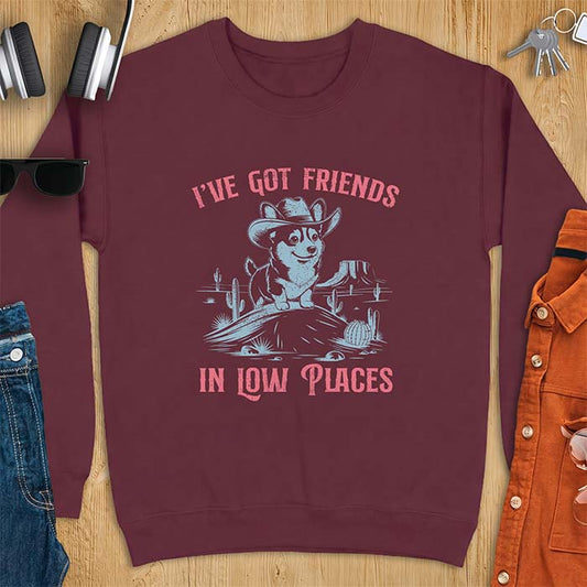 Maroon sweatshirt with a western graphic of a corgi puppy in a cowboy hat standing on a desert rock surrounded by cacti and a mesa, with the text I've Got Friends In Low Places printed above and below it.
