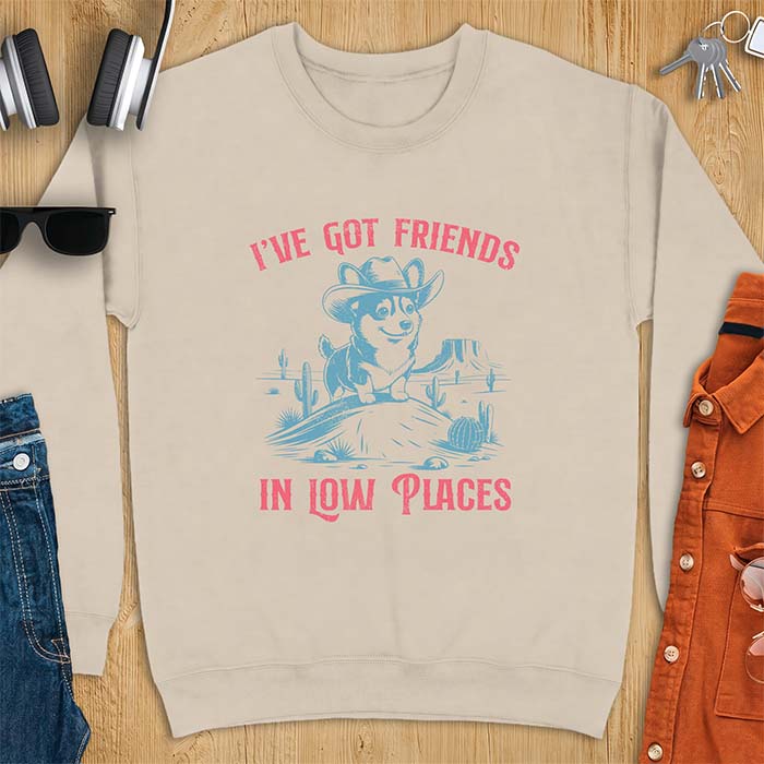 Sand sweatshirt with a western graphic of a corgi puppy in a cowboy hat standing on a desert rock surrounded by cacti and a mesa, with the text I've Got Friends In Low Places printed above and below it.