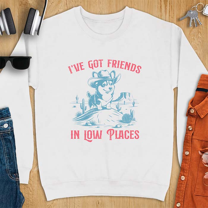 White sweatshirt with a western graphic of a corgi puppy in a cowboy hat standing on a desert rock surrounded by cacti and a mesa, with the text I've Got Friends In Low Places printed above and below it.