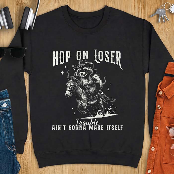 Black sweatshirt with a western graphic of a raccoon in a cowboy hat riding a donkey and an opossum riding along behind him under a scatter of stars and the text Hop On Loser, Trouble Ain't Gonna Make Itself