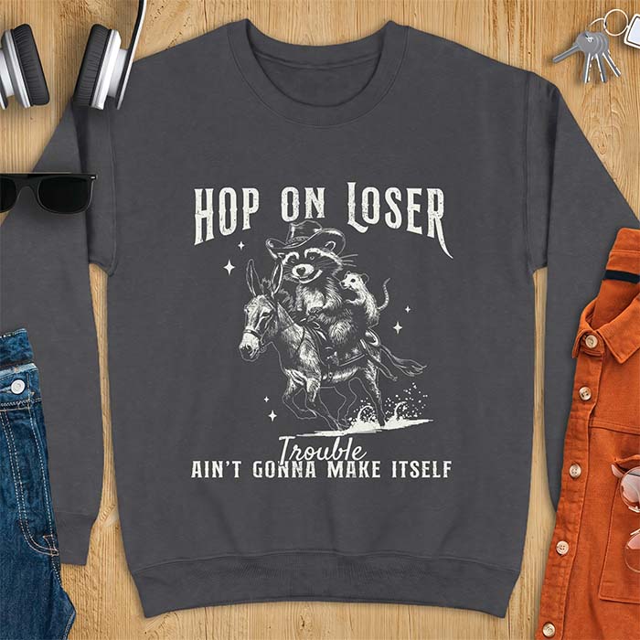 Dark Heather sweatshirt with a western graphic of a raccoon in a cowboy hat riding a donkey and an opossum riding along behind him under a scatter of stars and the text Hop On Loser, Trouble Ain't Gonna Make Itself