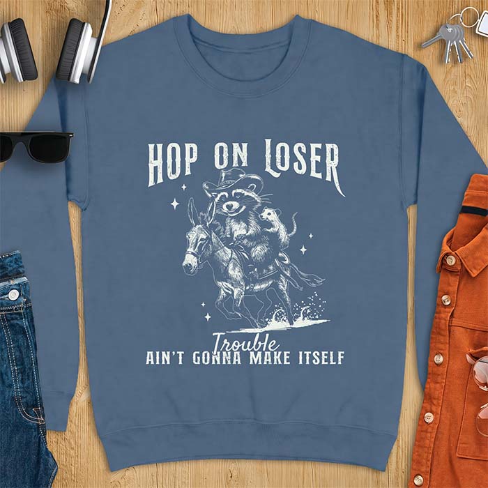 Indigo Blue sweatshirt with a western graphic of a raccoon in a cowboy hat riding a donkey and an opossum riding along behind him under a scatter of stars and the text Hop On Loser, Trouble Ain't Gonna Make Itself