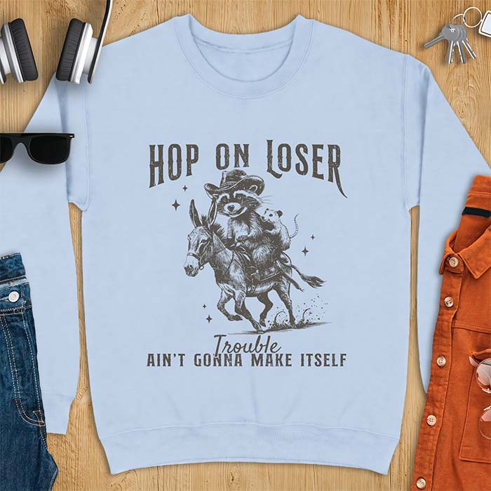 Light Blue sweatshirt with a western graphic of a raccoon in a cowboy hat riding a donkey and an opossum riding along behind him under a scatter of stars and the text Hop On Loser, Trouble Ain't Gonna Make Itself