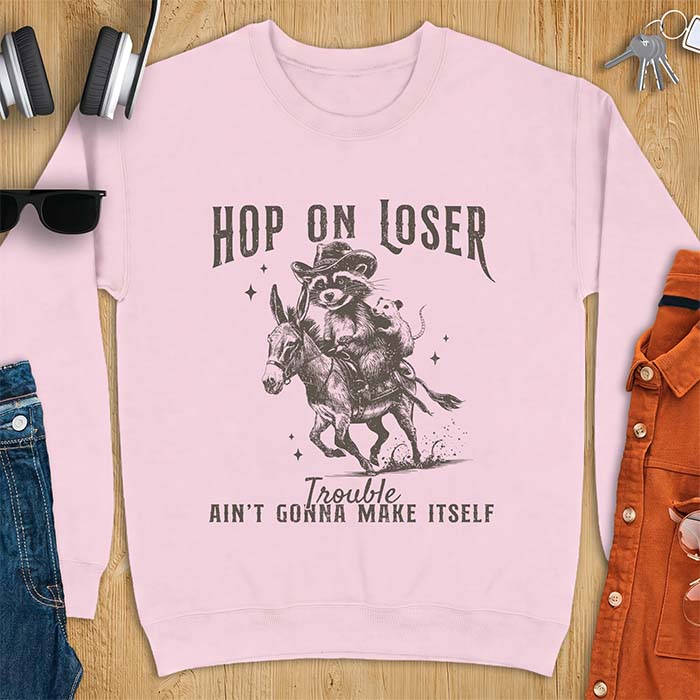 Light Pink sweatshirt with a western graphic of a raccoon in a cowboy hat riding a donkey and an opossum riding along behind him under a scatter of stars and the text Hop On Loser, Trouble Ain't Gonna Make Itself