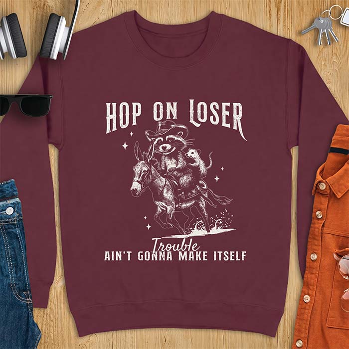 Maroon sweatshirt with a western graphic of a raccoon in a cowboy hat riding a donkey and an opossum riding along behind him under a scatter of stars and the text Hop On Loser, Trouble Ain't Gonna Make Itself