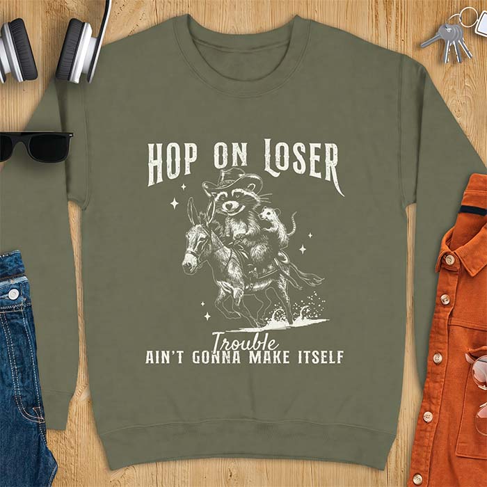 Military Green sweatshirt with a western graphic of a raccoon in a cowboy hat riding a donkey and an opossum riding along behind him under a scatter of stars and the text Hop On Loser, Trouble Ain't Gonna Make Itself