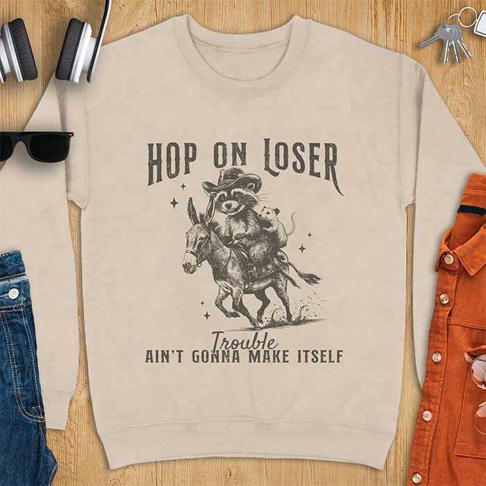Sand sweatshirt with a western graphic of a raccoon in a cowboy hat riding a donkey and an opossum riding along behind him under a scatter of stars and the text Hop On Loser, Trouble Ain't Gonna Make Itself