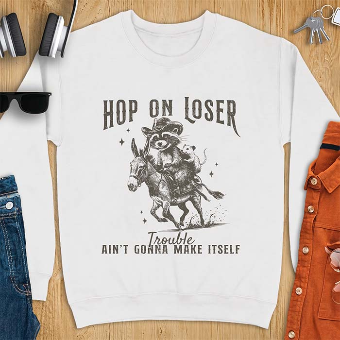 White sweatshirt with a western graphic of a raccoon in a cowboy hat riding a donkey and an opossum riding along behind him under a scatter of stars and the text Hop On Loser, Trouble Ain't Gonna Make Itself