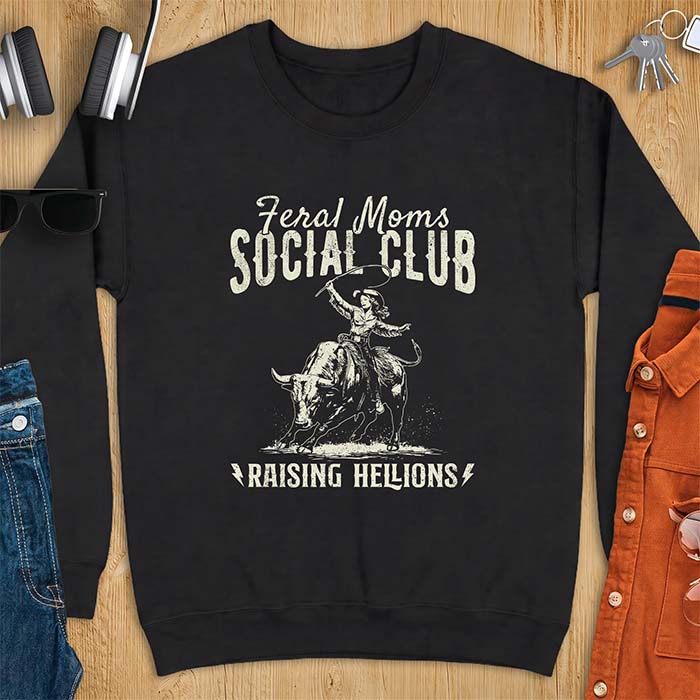 Black sweatshirt with a vintage western graphic of a cowgirl riding a bucking bull and swinging a whip, with the text Feral Moms Social Club above and Raising Hellions below
