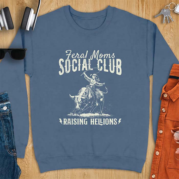 Indigo blue sweatshirt with a vintage western graphic of a cowgirl riding a bucking bull and swinging a whip, with the text Feral Moms Social Club above and Raising Hellions below