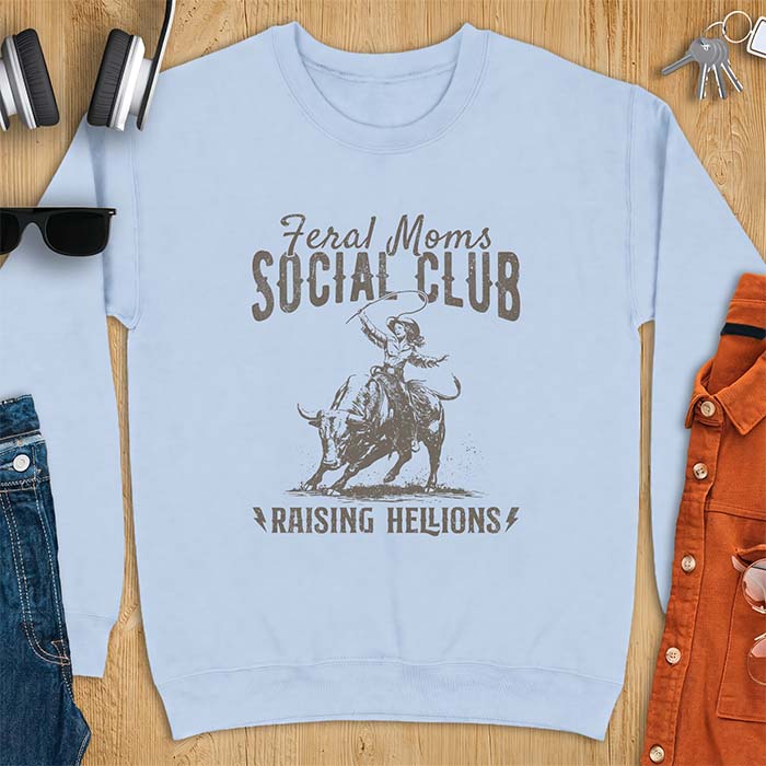 Light blue sweatshirt with a vintage western graphic of a cowgirl riding a bucking bull and swinging a whip, with the text Feral Moms Social Club above and Raising Hellions below