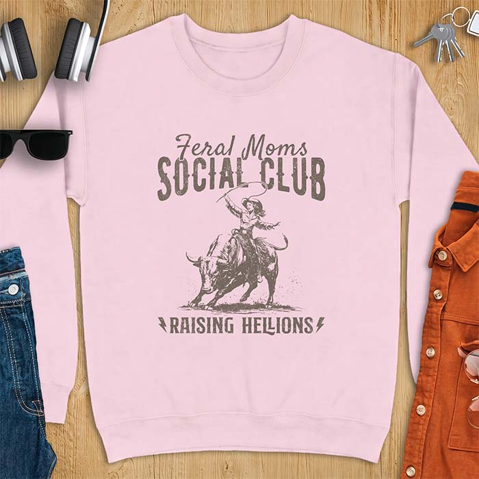 Light pink sweatshirt with a vintage western graphic of a cowgirl riding a bucking bull and swinging a whip, with the text Feral Moms Social Club above and Raising Hellions below
