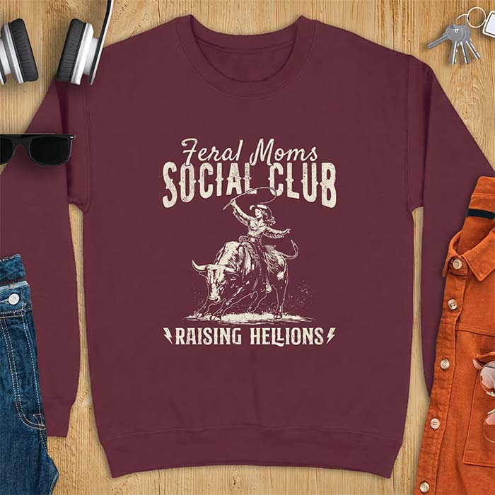 Maroon sweatshirt with a vintage western graphic of a cowgirl riding a bucking bull and swinging a whip, with the text Feral Moms Social Club above and Raising Hellions below