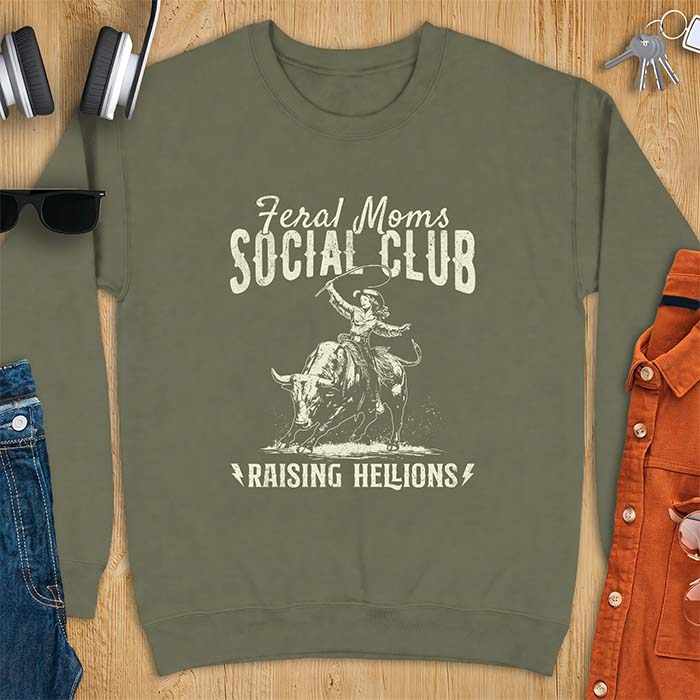 Military green sweatshirt with a vintage western graphic of a cowgirl riding a bucking bull and swinging a whip, with the text Feral Moms Social Club above and Raising Hellions below