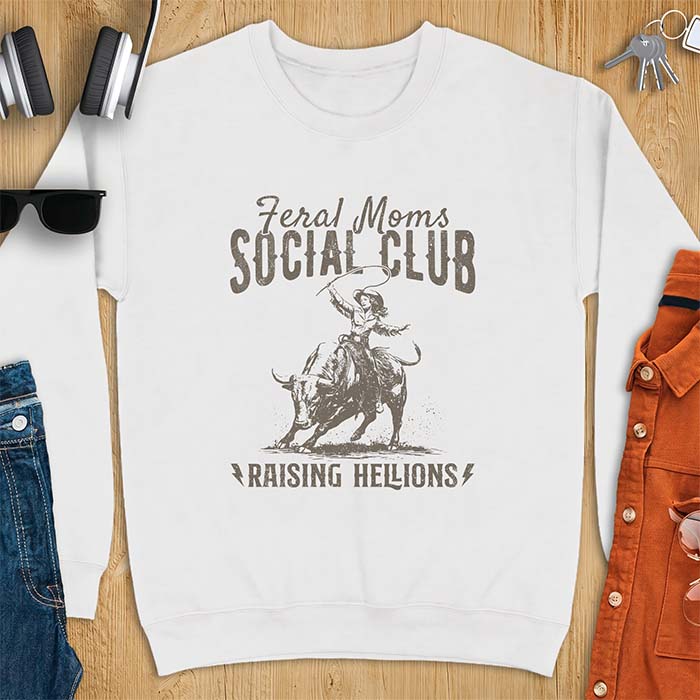 White sweatshirt with a vintage western graphic of a cowgirl riding a bucking bull and swinging a whip, with the text Feral Moms Social Club above and Raising Hellions below