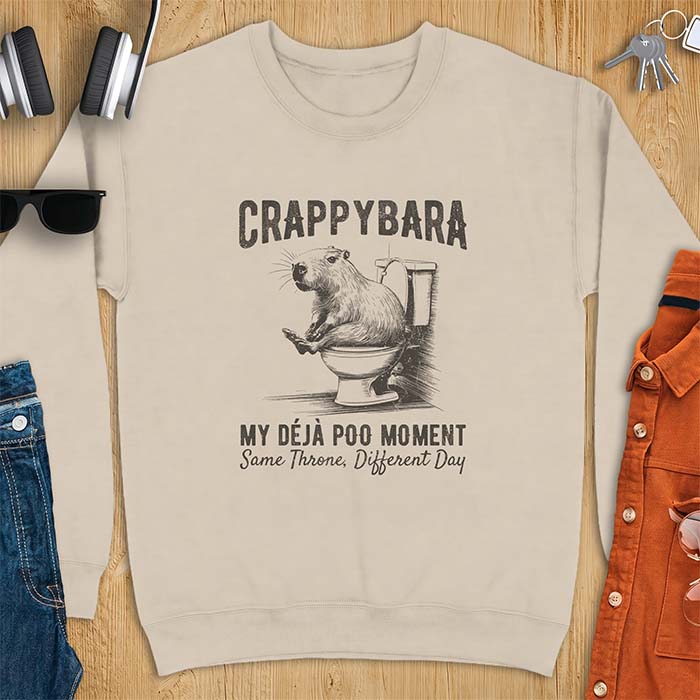 Sand sweatshirt with a toilet humor graphic of a capybara sitting on an old-fashioned toilet, printed with the words Crappybara, My Deja Poo Moment, Same Throne, Different Day.