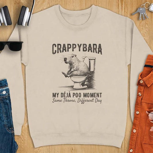 Sand sweatshirt with a toilet humor graphic of a capybara sitting on an old-fashioned toilet, printed with the words Crappybara, My Deja Poo Moment, Same Throne, Different Day.