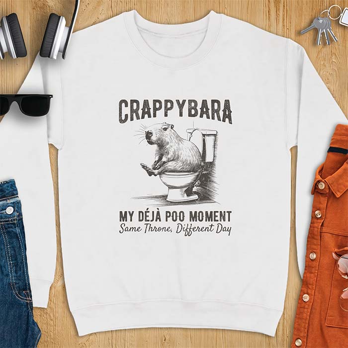 White sweatshirt with a toilet humor graphic of a capybara sitting on an old-fashioned toilet, printed with the words Crappybara, My Deja Poo Moment, Same Throne, Different Day.