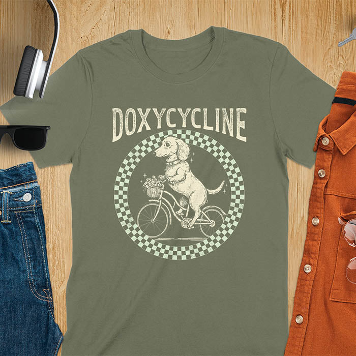 Military Green Dachshund t-shirt with 'DOXYCYCLINE' text and cute wiener dog riding bicycle in checkered circle design