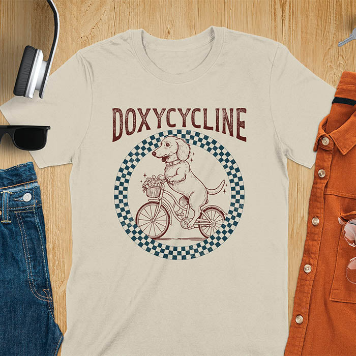 Sand color Dachshund t-shirt with 'DOXYCYCLINE' text and cute wiener dog riding bicycle in checkered circle design