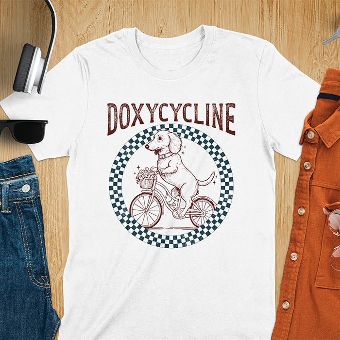 White Dachshund t-shirt with 'DOXYCYCLINE' text and cute wiener dog riding bicycle in checkered circle design