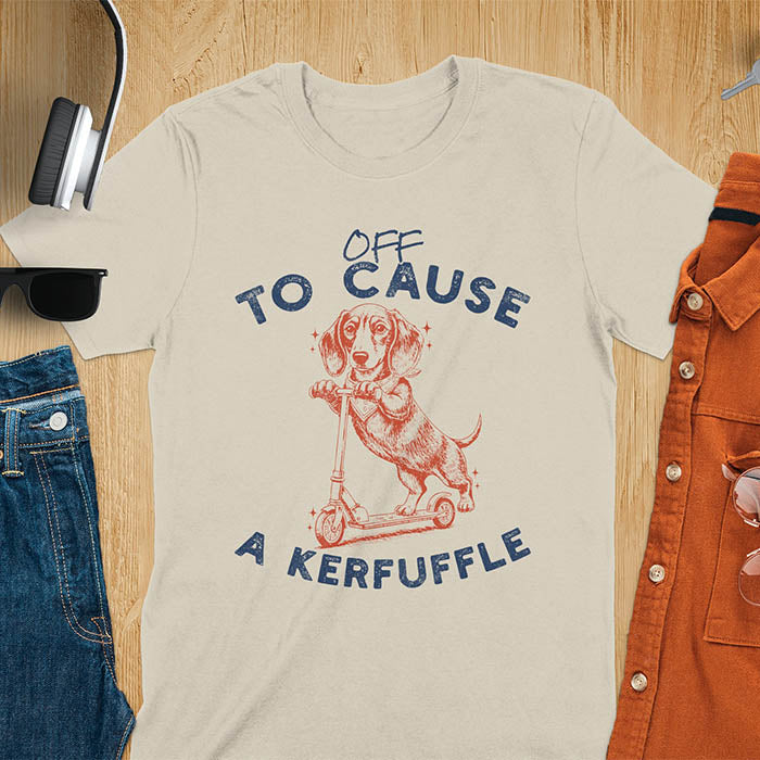 Dachshund on kick scooter illustrated graphic with Off To Cause A Kerfuffle text on sand color t-shirt
