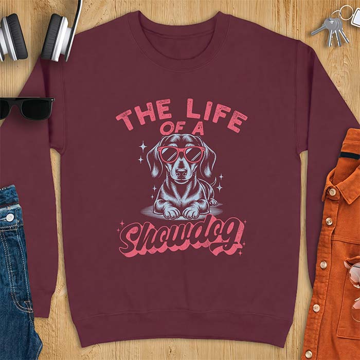 Maroon sweatshirt with a graphic of a Dachshund wearing red sunglasses lying down surrounded by sparkles, with the text The Life Of A Showdog