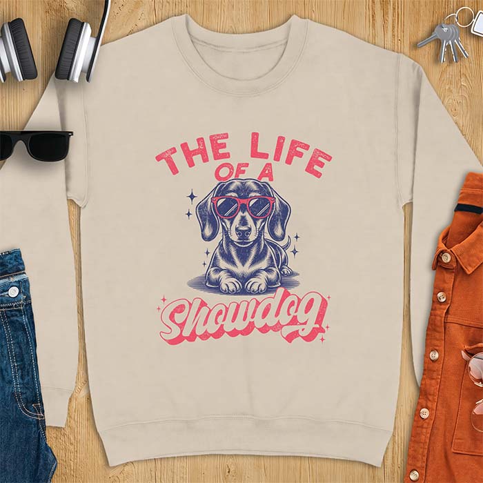 Sand sweatshirt with a graphic of a Dachshund wearing red sunglasses lying down surrounded by sparkles, with the text The Life Of A Showdog