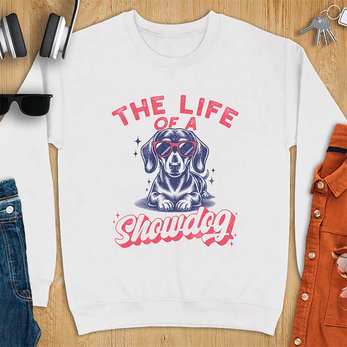 White sweatshirt with a graphic of a Dachshund wearing red sunglasses lying down surrounded by sparkles, with the text The Life Of A Showdog