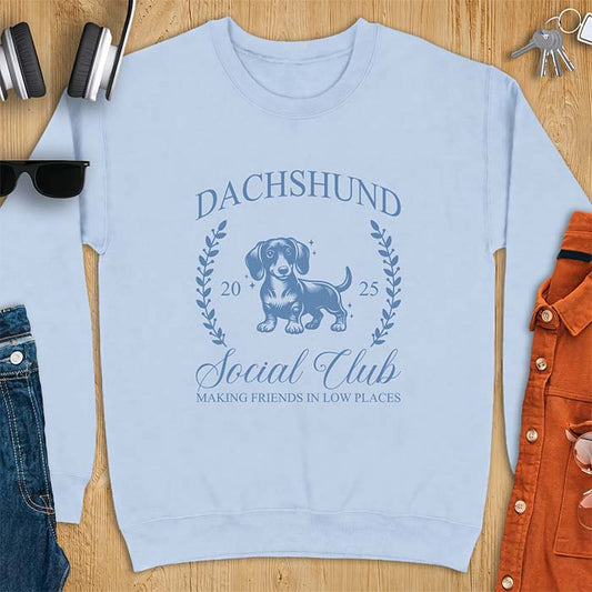 Light Blue sweatshirt with a vintage crest graphic of a dachshund standing inside a laurel wreath alongside the text Dachshund, Social Club, 2025, Making Friends In Low Places.