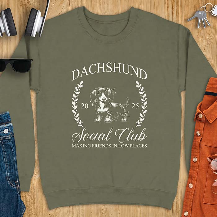 Military Green sweatshirt with a vintage crest graphic of a dachshund standing inside a laurel wreath alongside the text Dachshund, Social Club, 2025, Making Friends In Low Places.