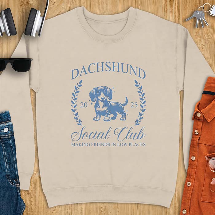 Sand sweatshirt with a vintage crest graphic of a dachshund standing inside a laurel wreath alongside the text Dachshund, Social Club, 2025, Making Friends In Low Places.
