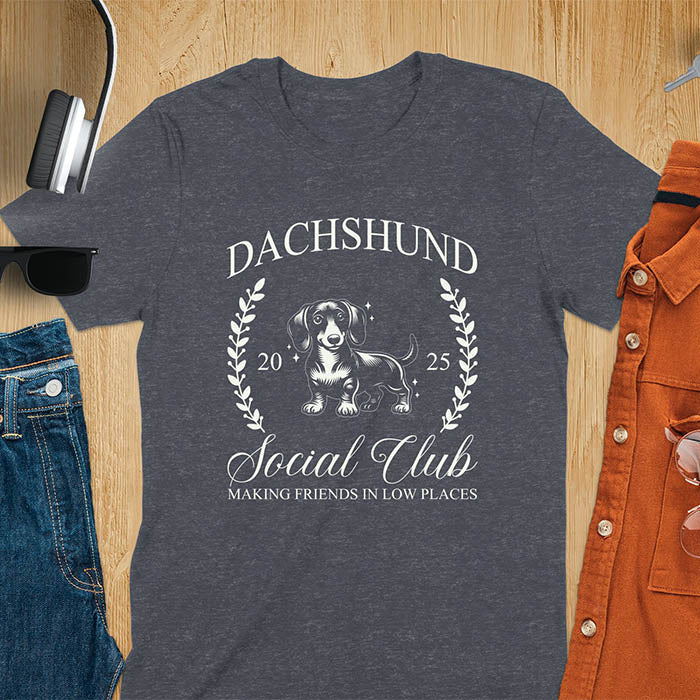 Dark heather T-shirt with a playful dachshund graphic, laurel branches, and text reading "Dachshund Social Club," "EST 2024," and "Making Friends in Low Places." Vintage-inspired design perfect for dog lovers.