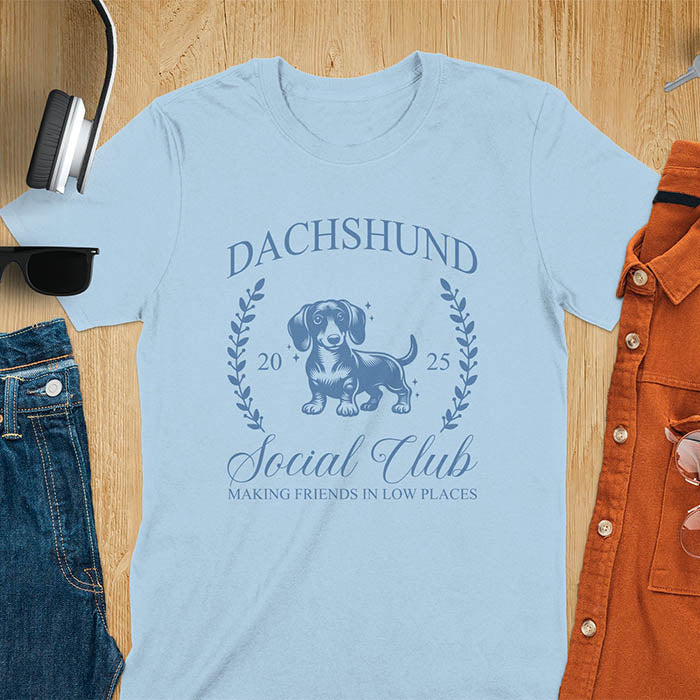Light blue T-shirt with a playful dachshund graphic, laurel branches, and text reading "Dachshund Social Club," "EST 2024," and "Making Friends in Low Places." Vintage-inspired design perfect for dog lovers.