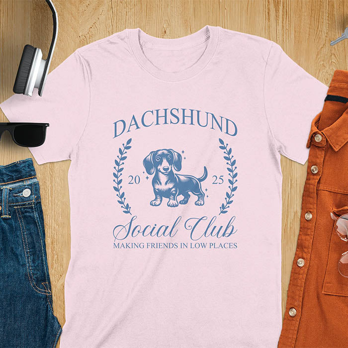 Light pink T-shirt with a playful dachshund graphic, laurel branches, and text reading "Dachshund Social Club," "EST 2024," and "Making Friends in Low Places." Vintage-inspired design perfect for dog lovers.