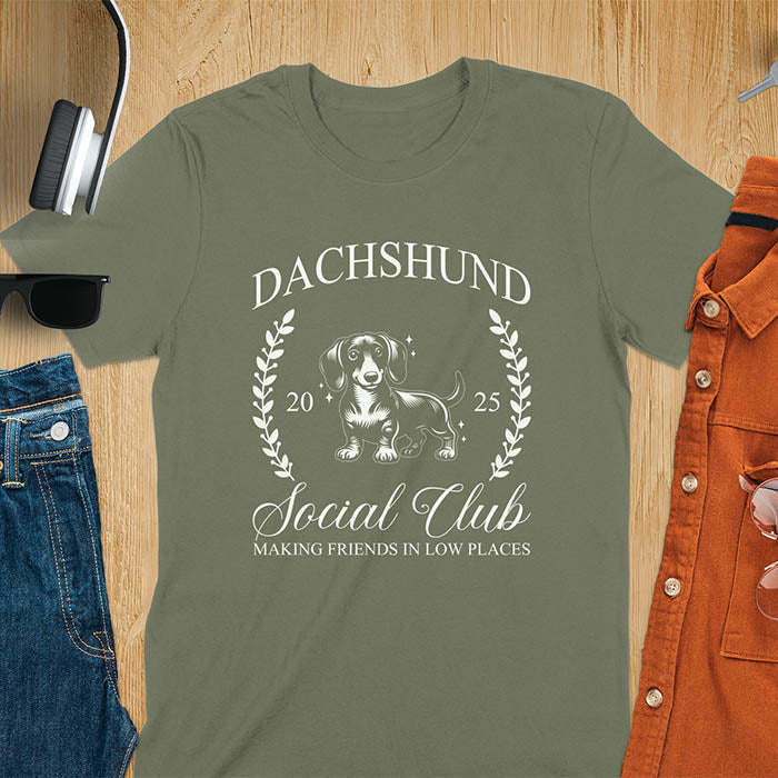 Military green T-shirt with a playful dachshund graphic, laurel branches, and text reading "Dachshund Social Club," "EST 2024," and "Making Friends in Low Places." Vintage-inspired design perfect for dog lovers.