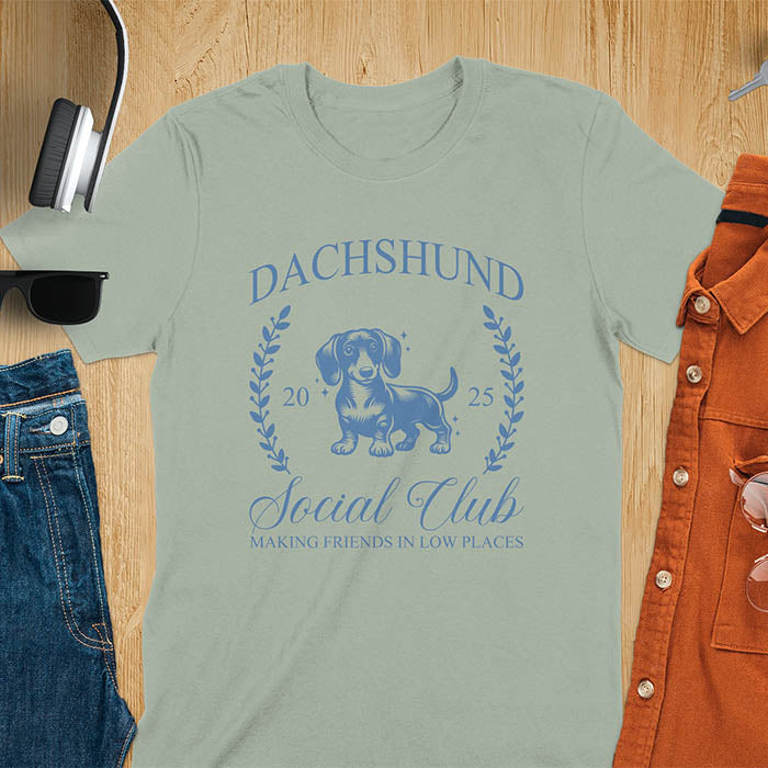 Sage green T-shirt with a playful dachshund graphic, laurel branches, and text reading "Dachshund Social Club," "EST 2024," and "Making Friends in Low Places." Vintage-inspired design perfect for dog lovers.
